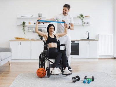Husband in sportswear helping mature woman in wheelchair lifting arms with stretching bands for muscle strength. Physiotherapy healthcare, medical trainer consulting disabled caucasian patient.