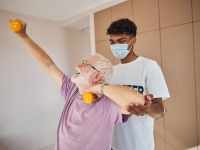 Pensioner performing a strength exercise with a pair of dumbbells assisted by his fitness trainer